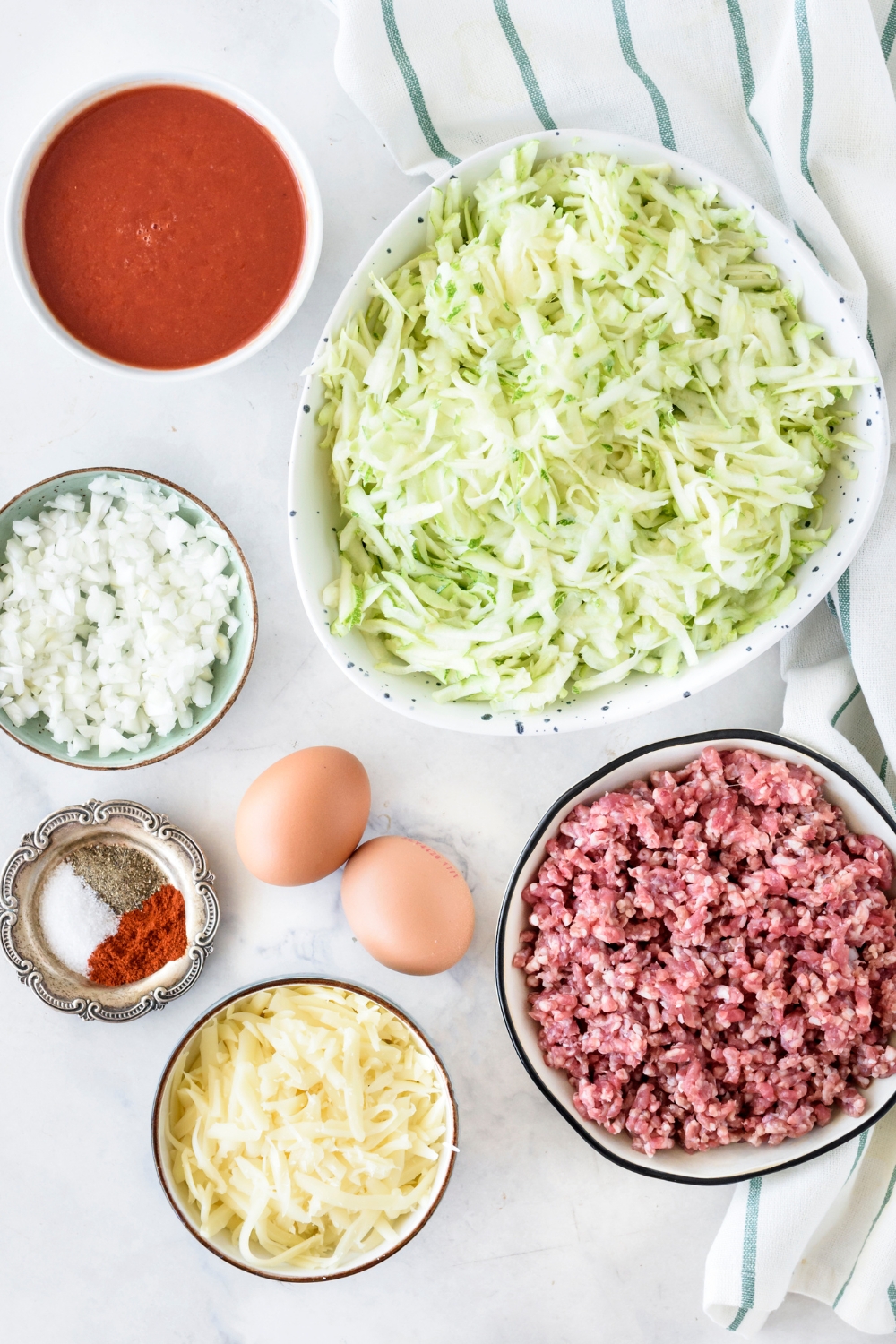 An assortment of ingredients including bowls of grated zucchini, raw ground beef, tomato sauce, diced onion, shredded cheese, spices, and two eggs.