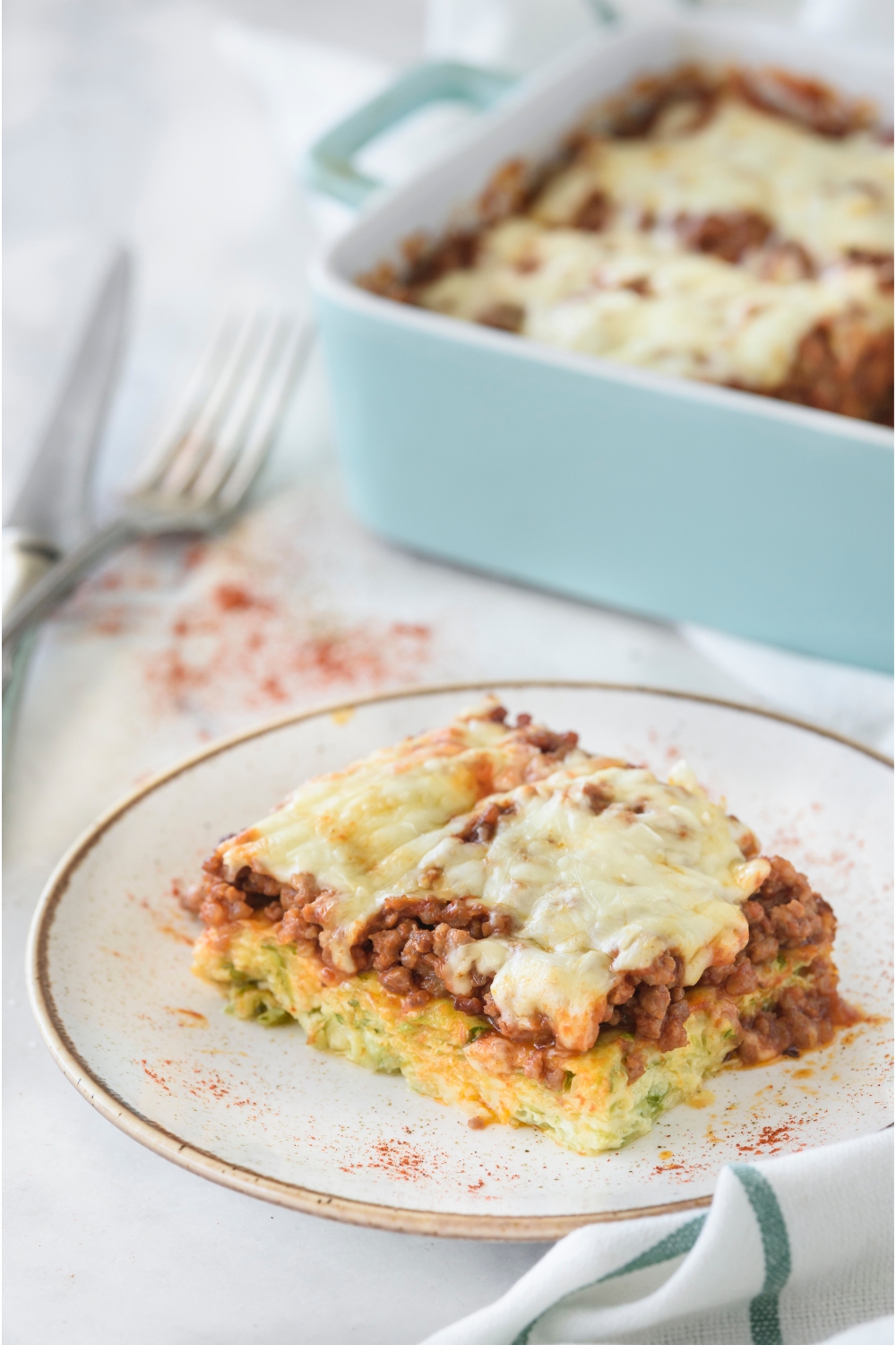 A square serving of zucchini pizza casserole topped with a layer of melted cheese and dusted with paprika. The rest of the casserole is in the background.