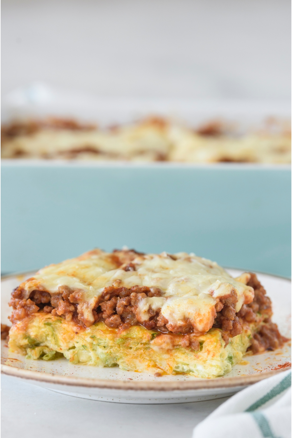 A square serving of zucchini pizza casserole topped with a layer of melted cheese. The rest of the casserole is in the background.