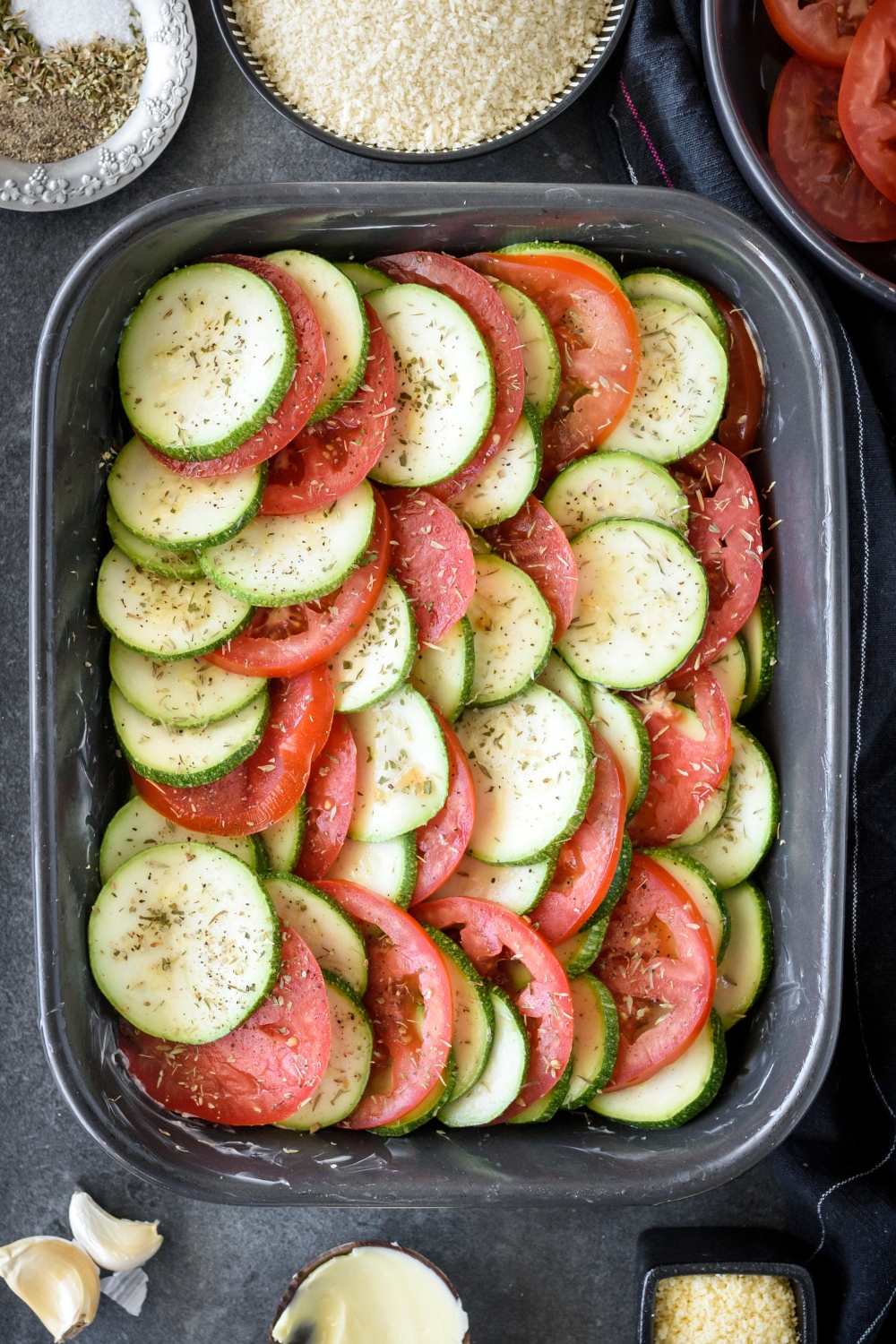 A baking pan with sliced zucchini and tomatoes placed in a layer seasoning with herbs and seasonings.
