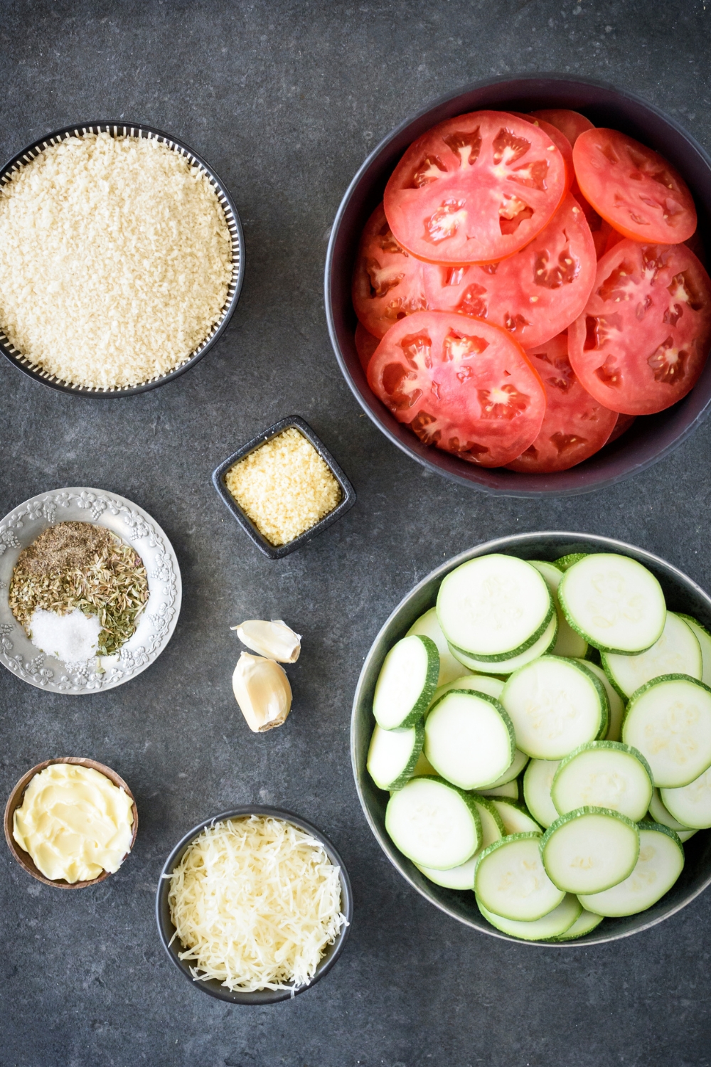 A countertop with sliced tomatoes, sliced zucchini, shredded cheese, butter, garlic, herbs, and seasonings in various bowls.