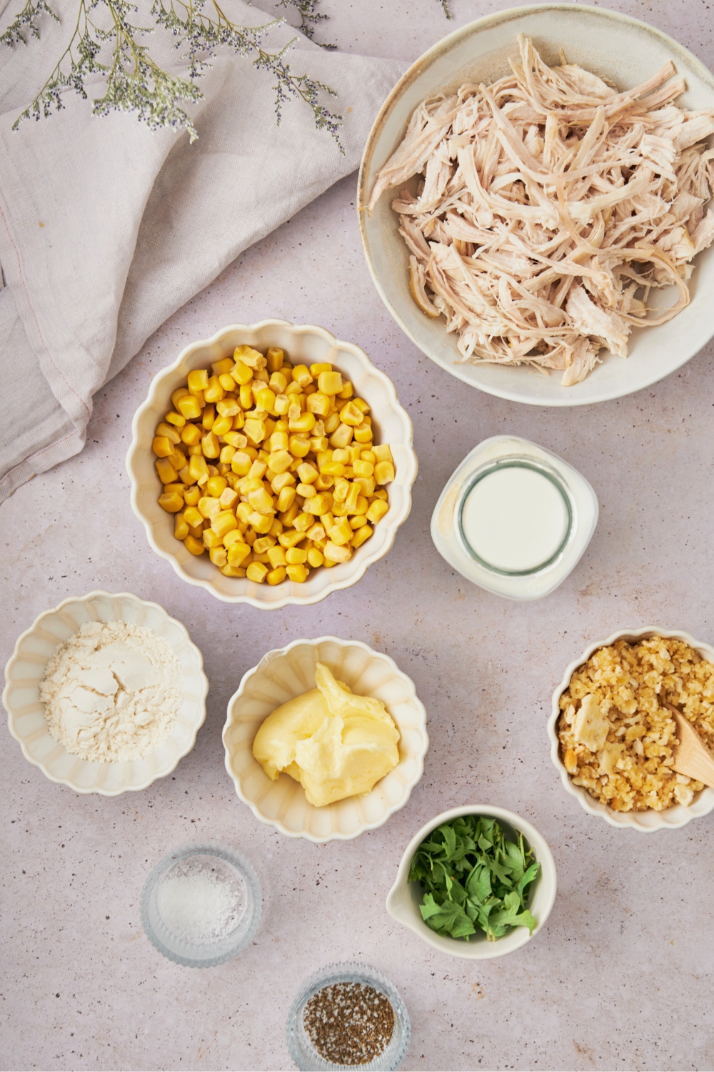 A bowl of shredded chicken, a bowl of corn, a bowl of flour, a bowl of butter, and a bowl of chicken all on a grey counter.