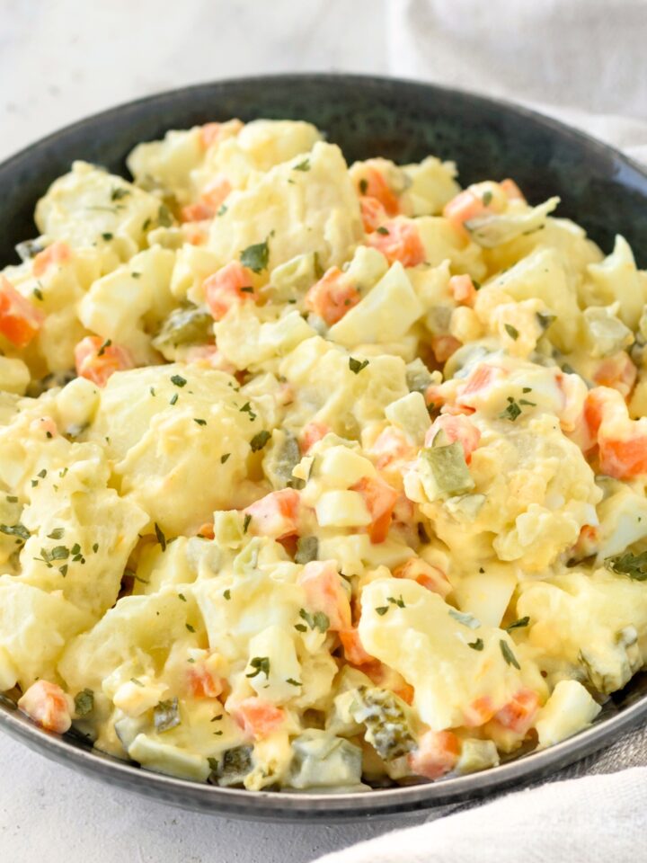 A bowl of potato salad with chunks of pickles and carrots. The potato salad is garnished with herbs.