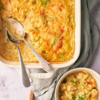 Part of a white casserole dish that has chicken and rice casserole in it. Two spoons are in the dish in part of where some of the casserole is missing. That scoop of casserole is in a white bowl in front of the casserole dish.