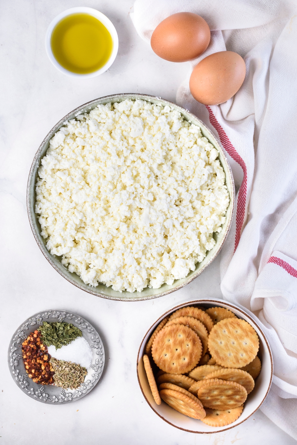 A countertop with ricotta cheese, crackers, seasonings and herbs, eggs, and olive oil.