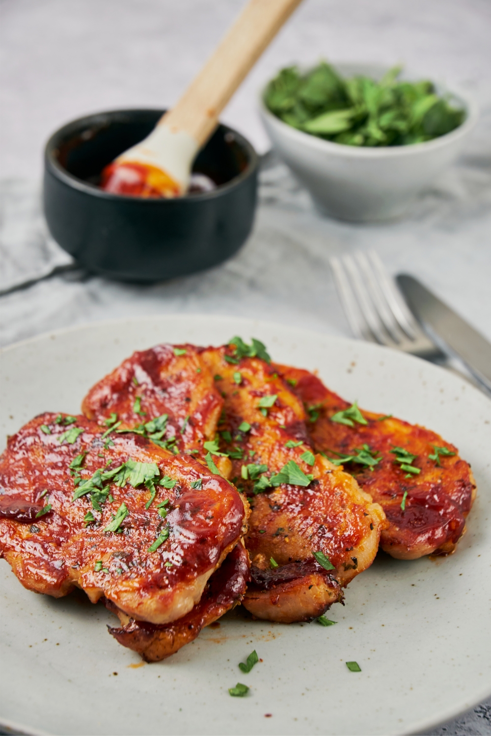 Four oven baked BBQ pork chops garnished with herbs on a white plate with a small bowl of extra sauce in the back.