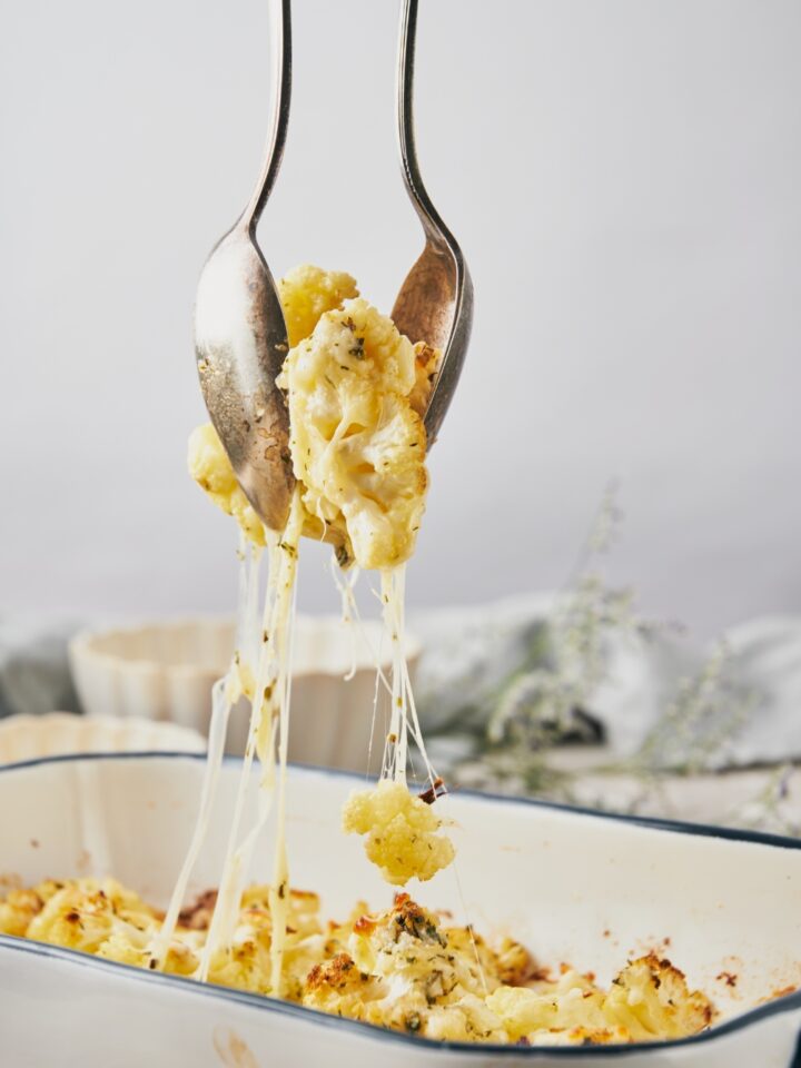 A casserole dish with two spoons scooping a serving of cheesy cauliflower.