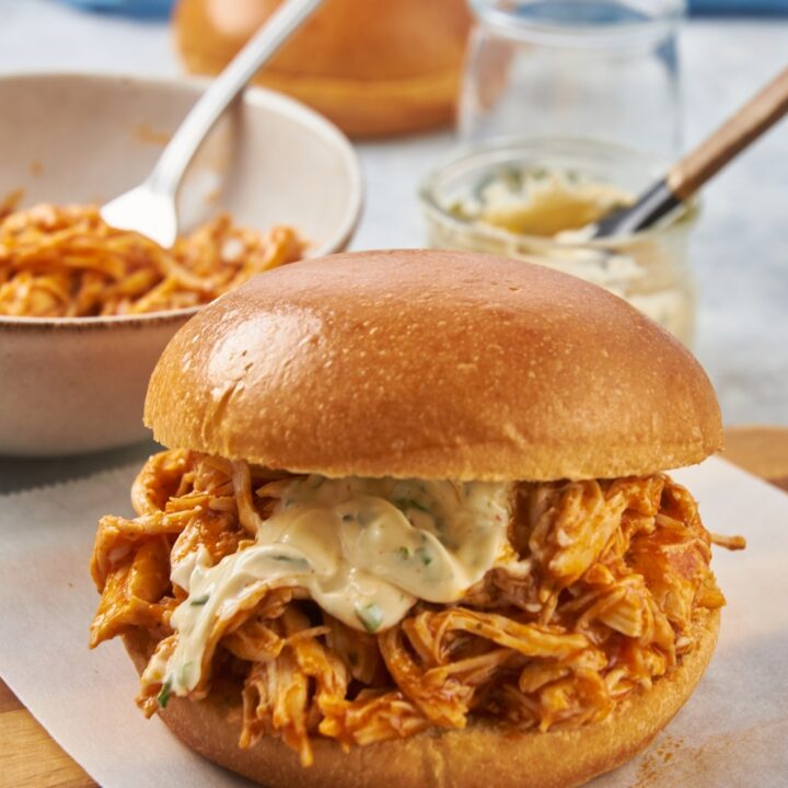 A shredded buffalo chicken sandwich with hamburger buns next to a bowl with more shredded chicken and a fork.