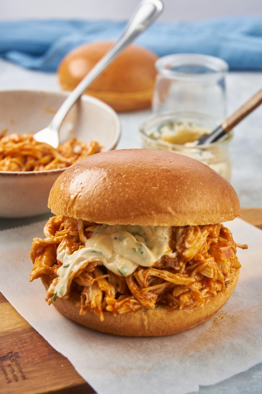 A shredded buffalo chicken sandwich with hamburger buns next to a bowl with more shredded chicken and a fork.