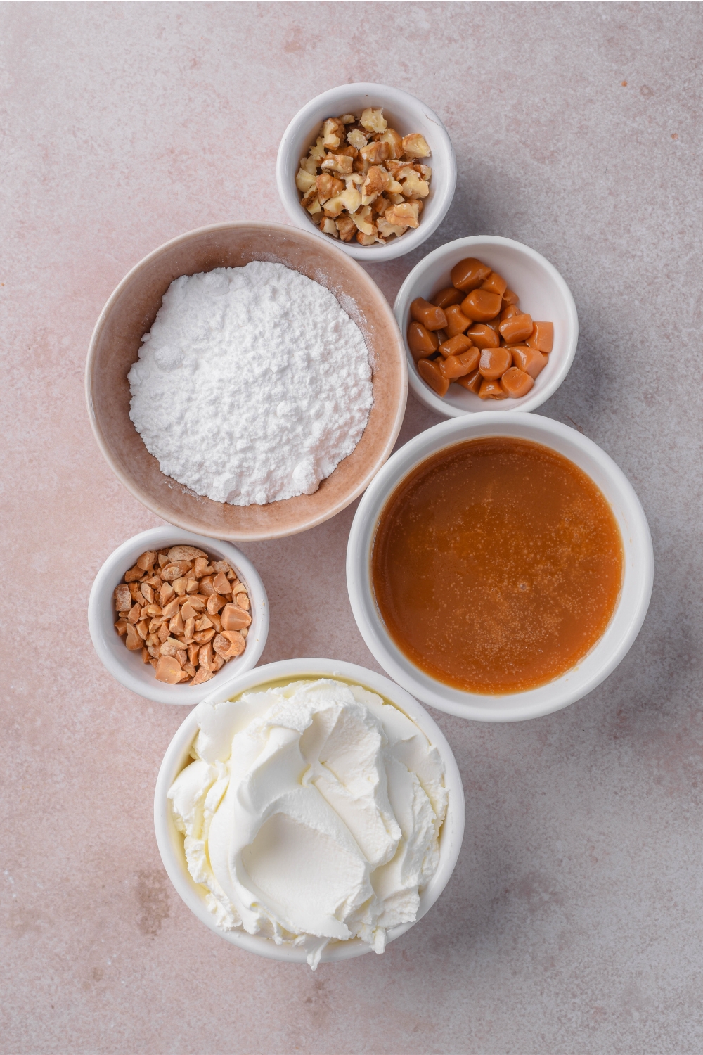 A bowl of toffee bits, a bowl of walnuts, a bowl of peanuts, a bowl of powdered sugar, a bowl of caramel, and a bowl of cream cheese.