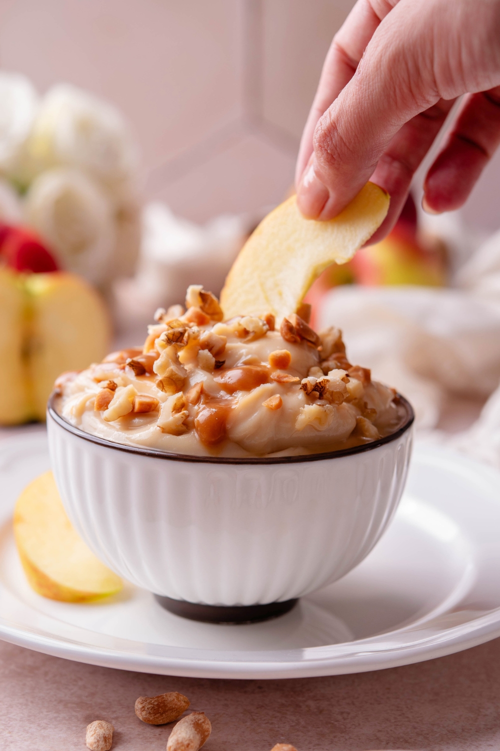 A hand holding an apple slice dipping it into a white bowl that's filled with caramel apple dip.