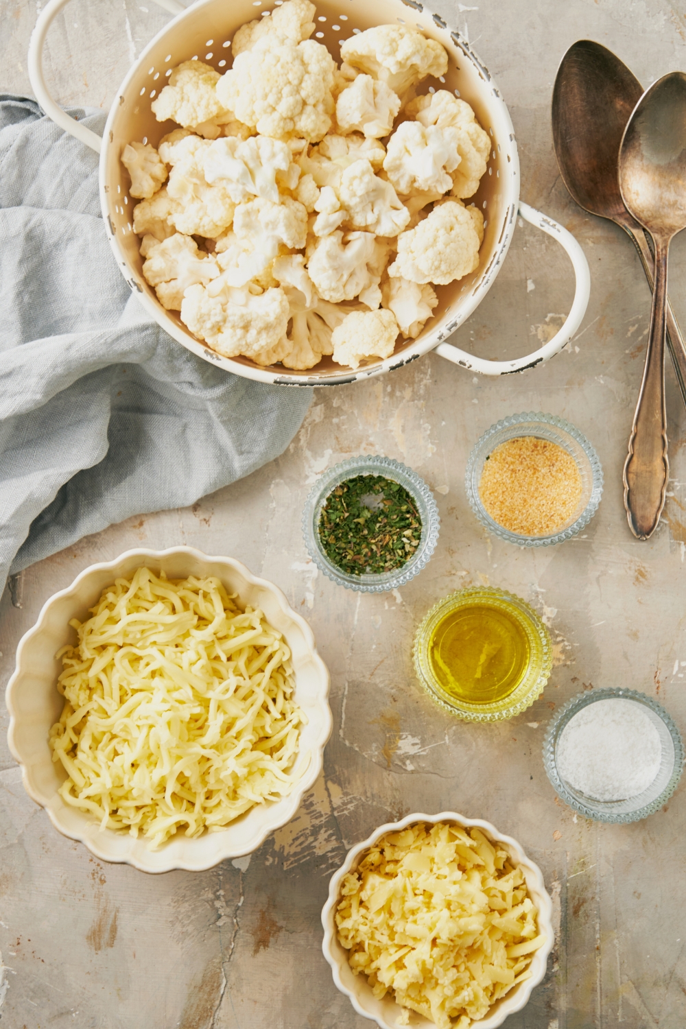 A countertop with cauliflower, shredded cheese, seasonings, oil, and salt in various bowls.