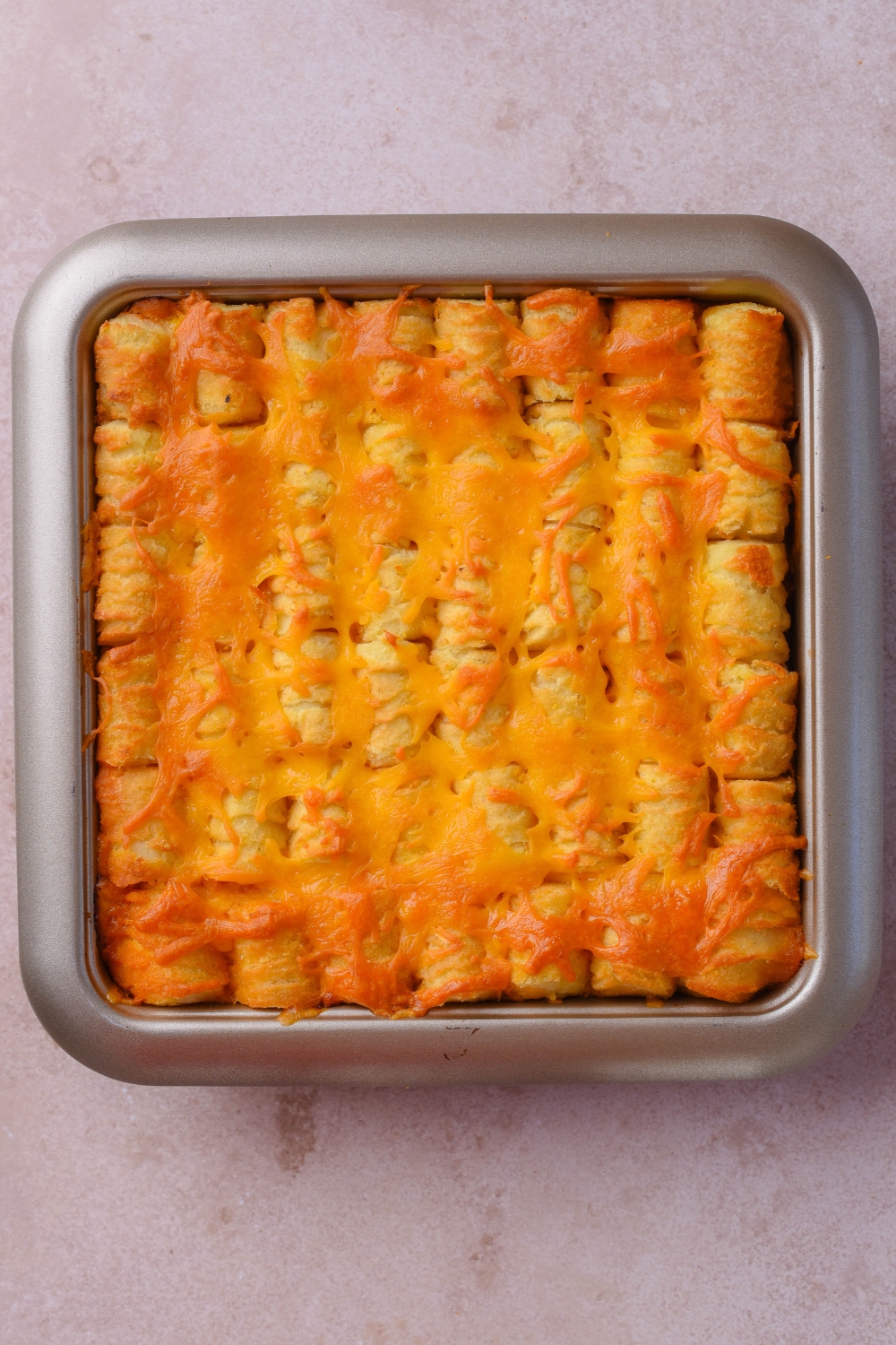 Melted cheese on top of tater tots layered in a square baking dish on a grey counter.