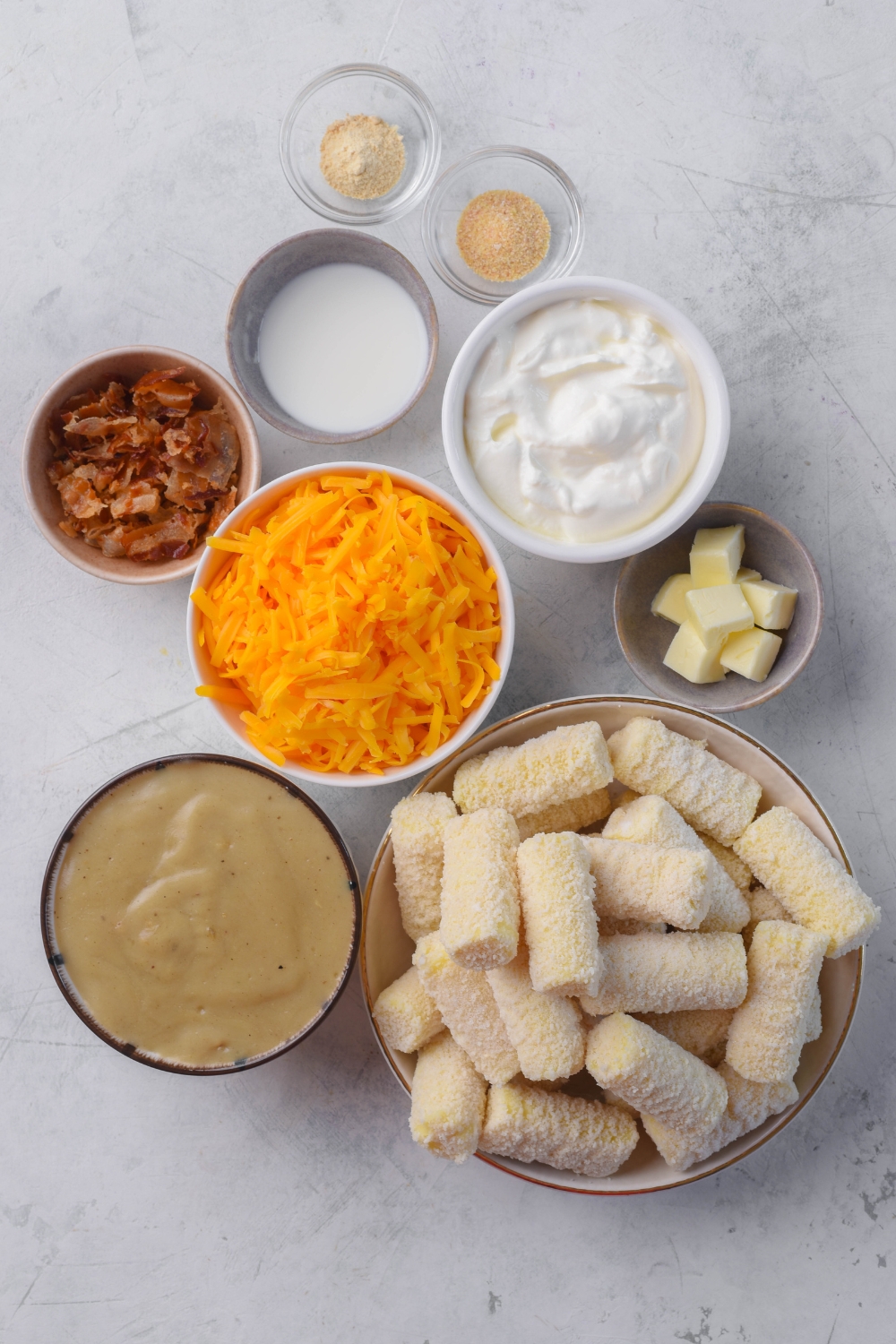 A countertop with sour cream, shredded cheese, condensed soup, bacon bits, tater tots, cubed butter, milk, onion powder, and garlic powder in separate bowls.