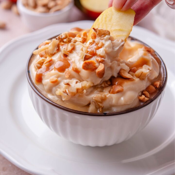 A hand dipping an apple slice into a caramel apple dip.