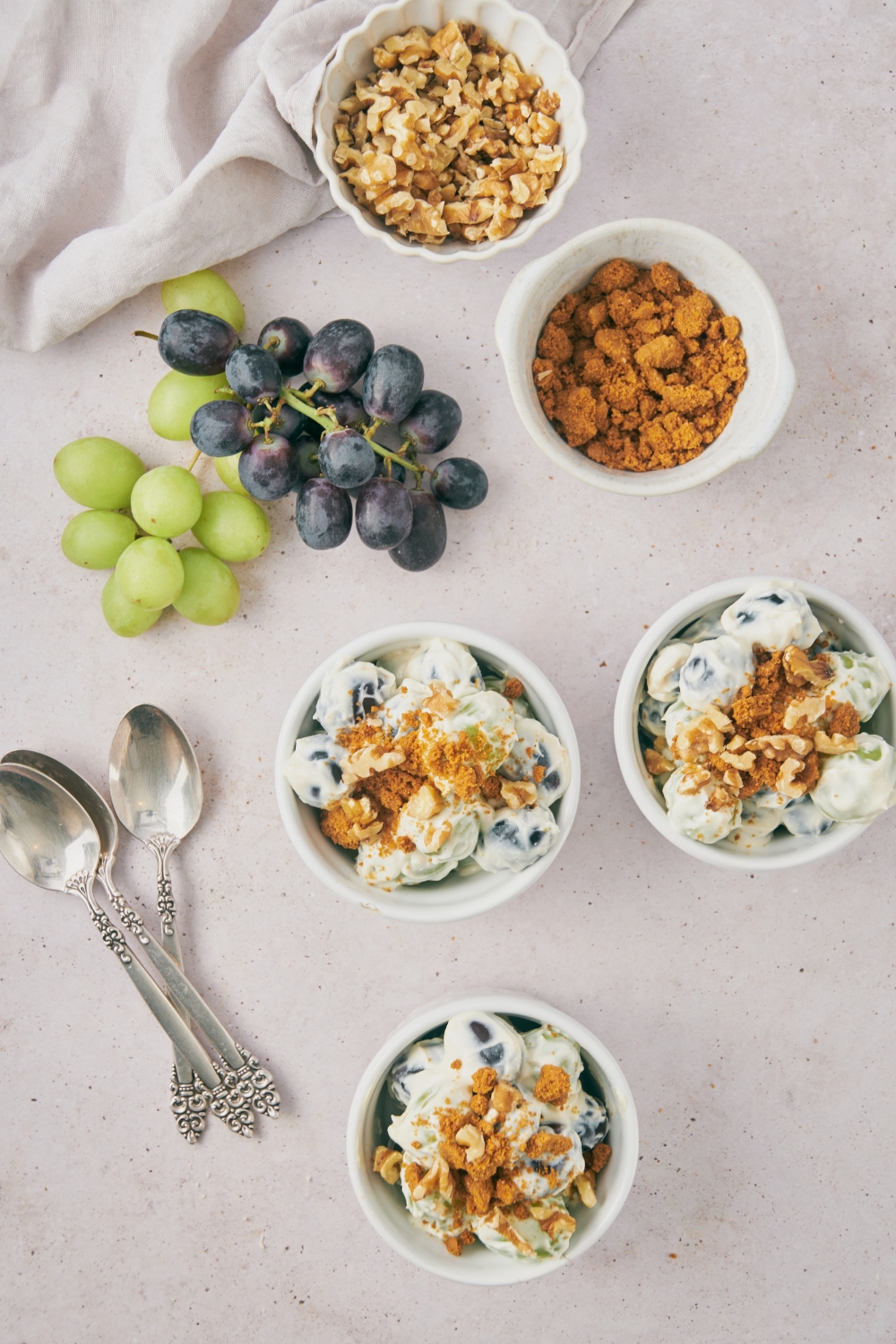 Three bowls of grape salad, a vine of purple grapes, a vine of green grapes, a bowl of cookie crumbs, and a bowl of crushed walnuts all on a grey counter.