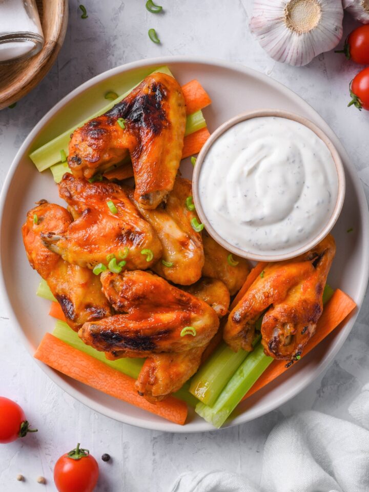 A bunch of chicken wings coated in hot sauce on top of a white plate with a cup of ranch dressing, carrot sticks, and celery sticks.