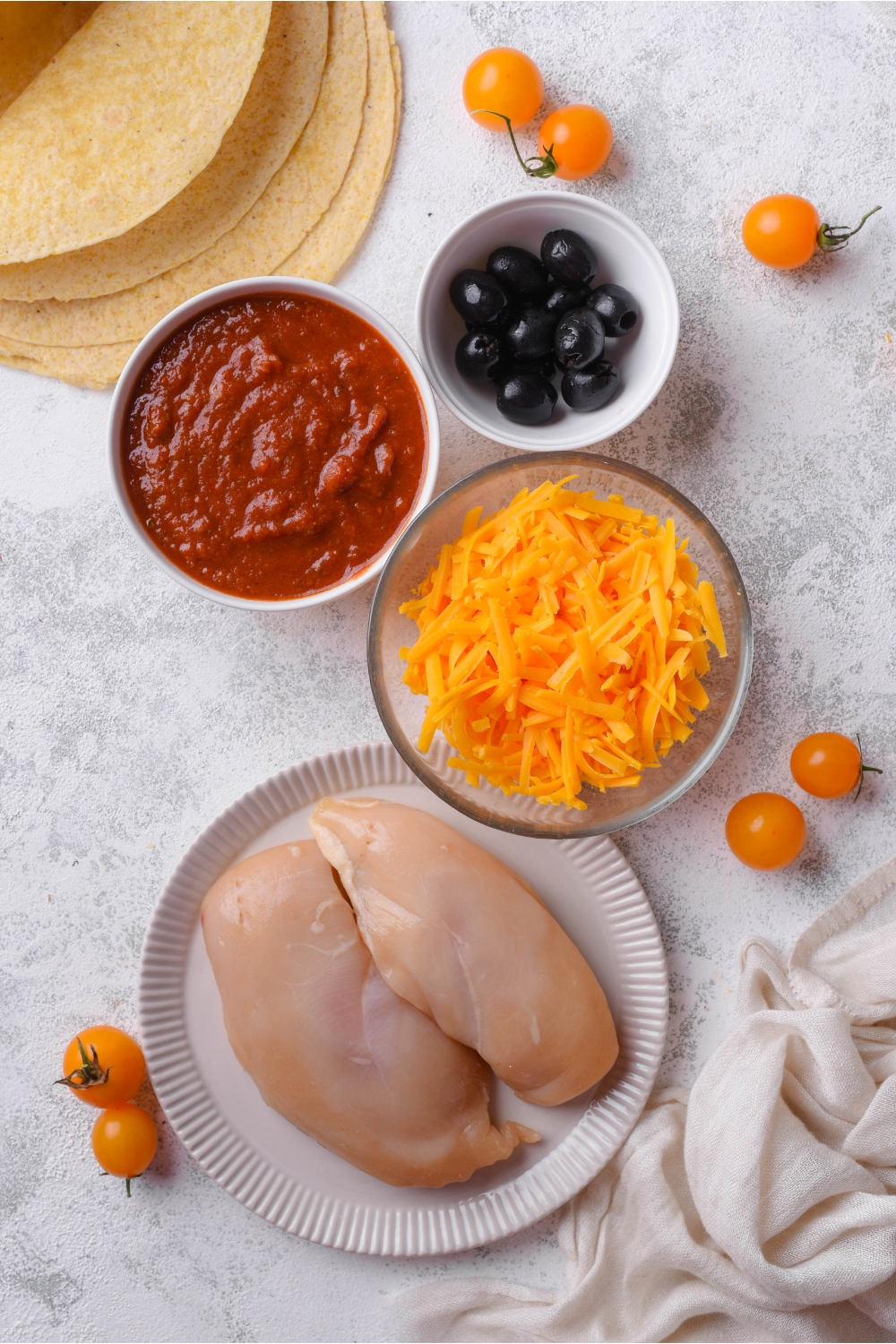 A countertop with chicken breasts, shredded cheese, enchilada sauce, olives, and corn tortillas in separate bowls.