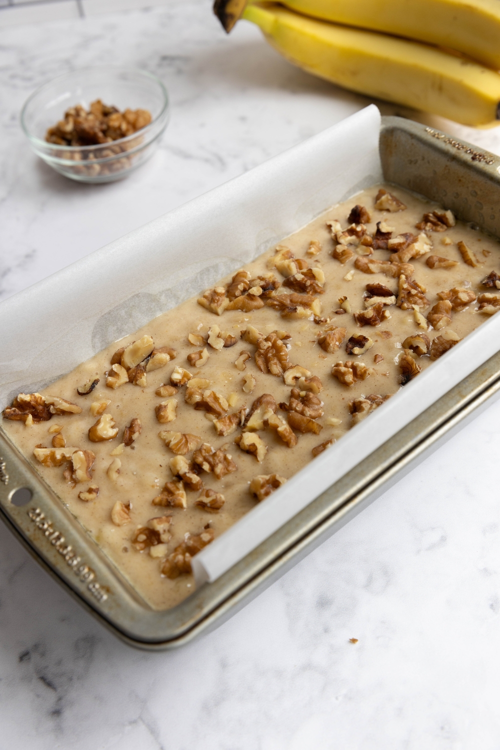 A loaf pan with banana bread batter topped with chopped walnuts.