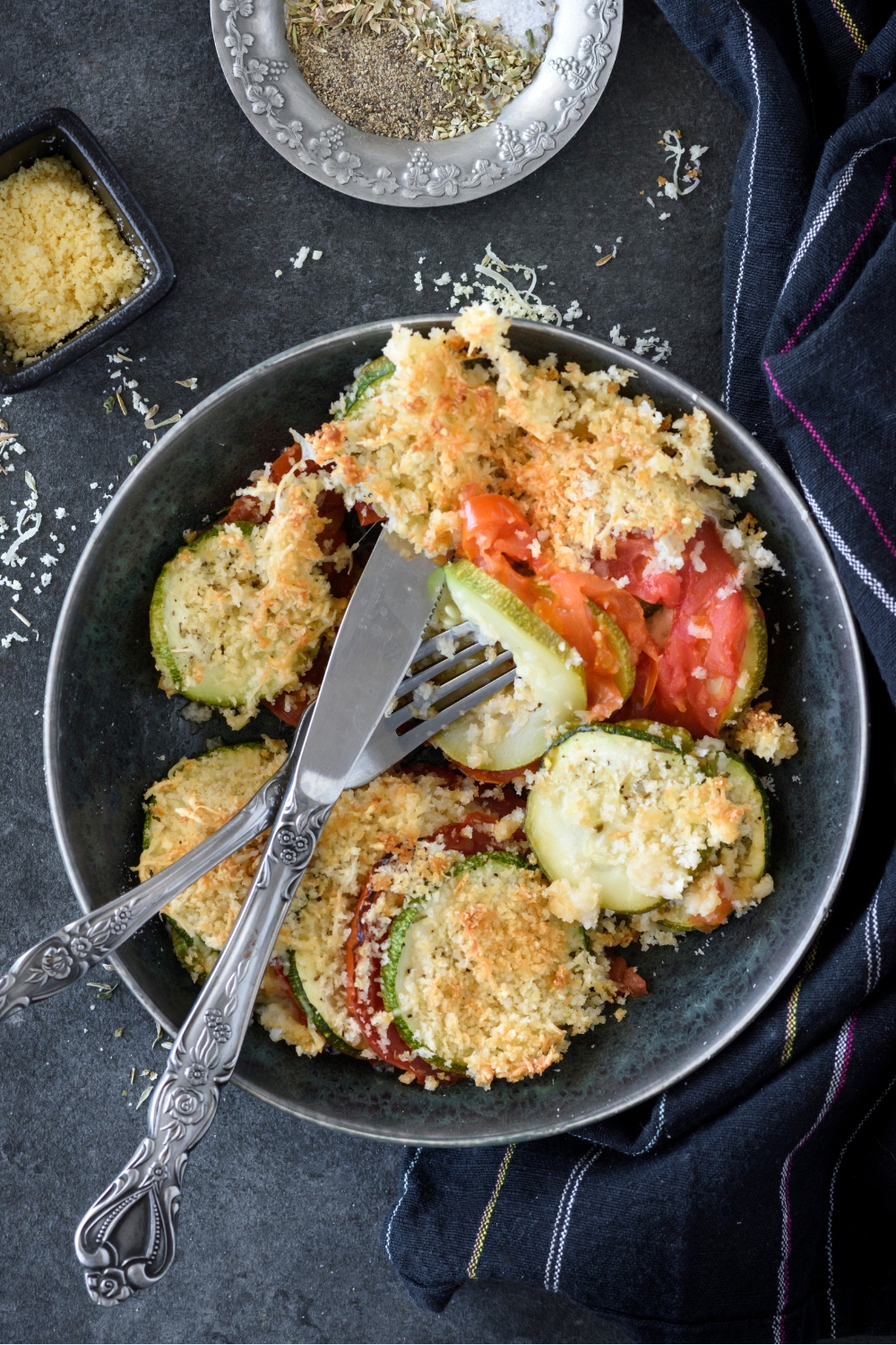 A bowl with zucchini tomato casserole and a fork and knife.
