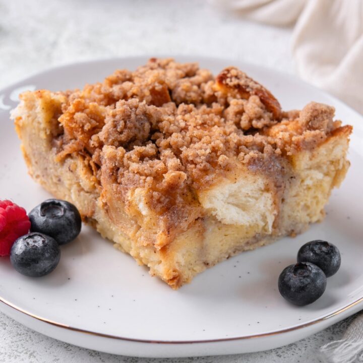 A square slice of brioche french toast casserole on a white plate with some blueberries on each side.