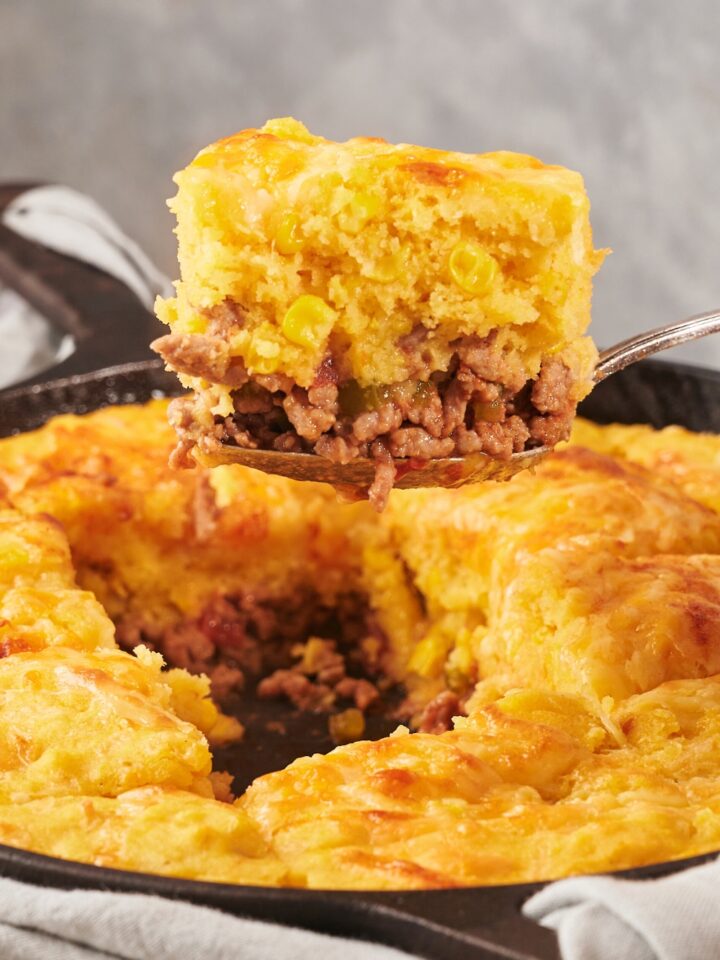A spoon holding a slice of mexican cornbread casserole over a cast iron skillet with the casserole.