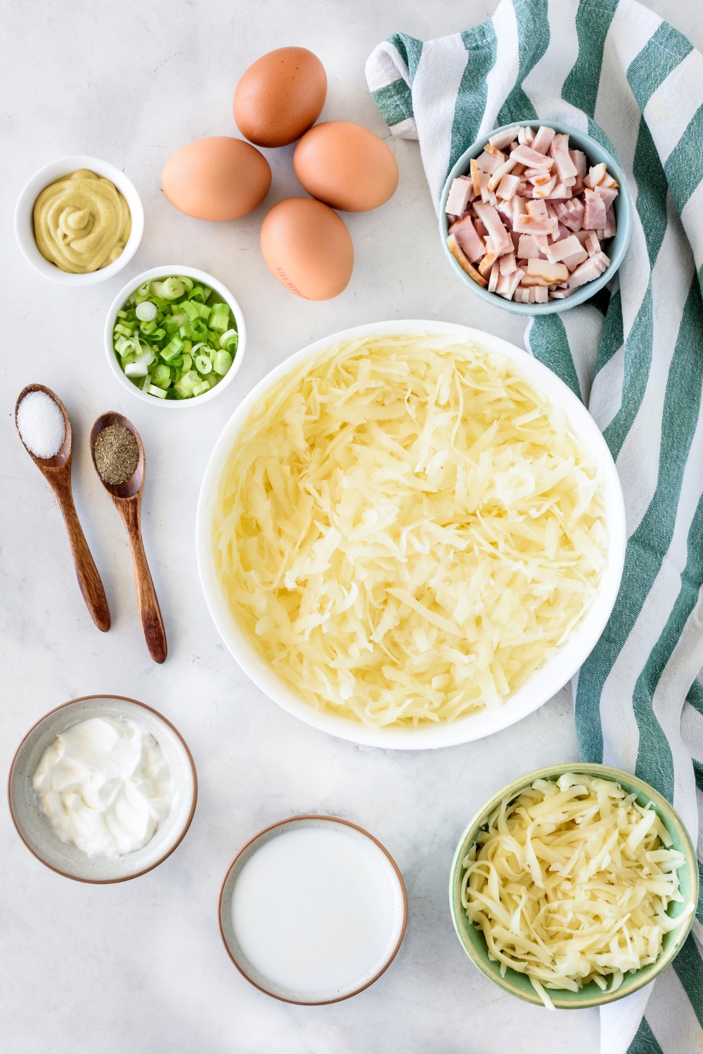 A countertop with hash browns, eggs, green onions, dijon mustard, diced bacon, salt, pepper, onions, cream, and shredded cheese.