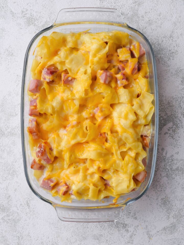 Melted cheddar cheese on top of egg noodles and diced ham in a glass casserole dish on top of a grey counter.