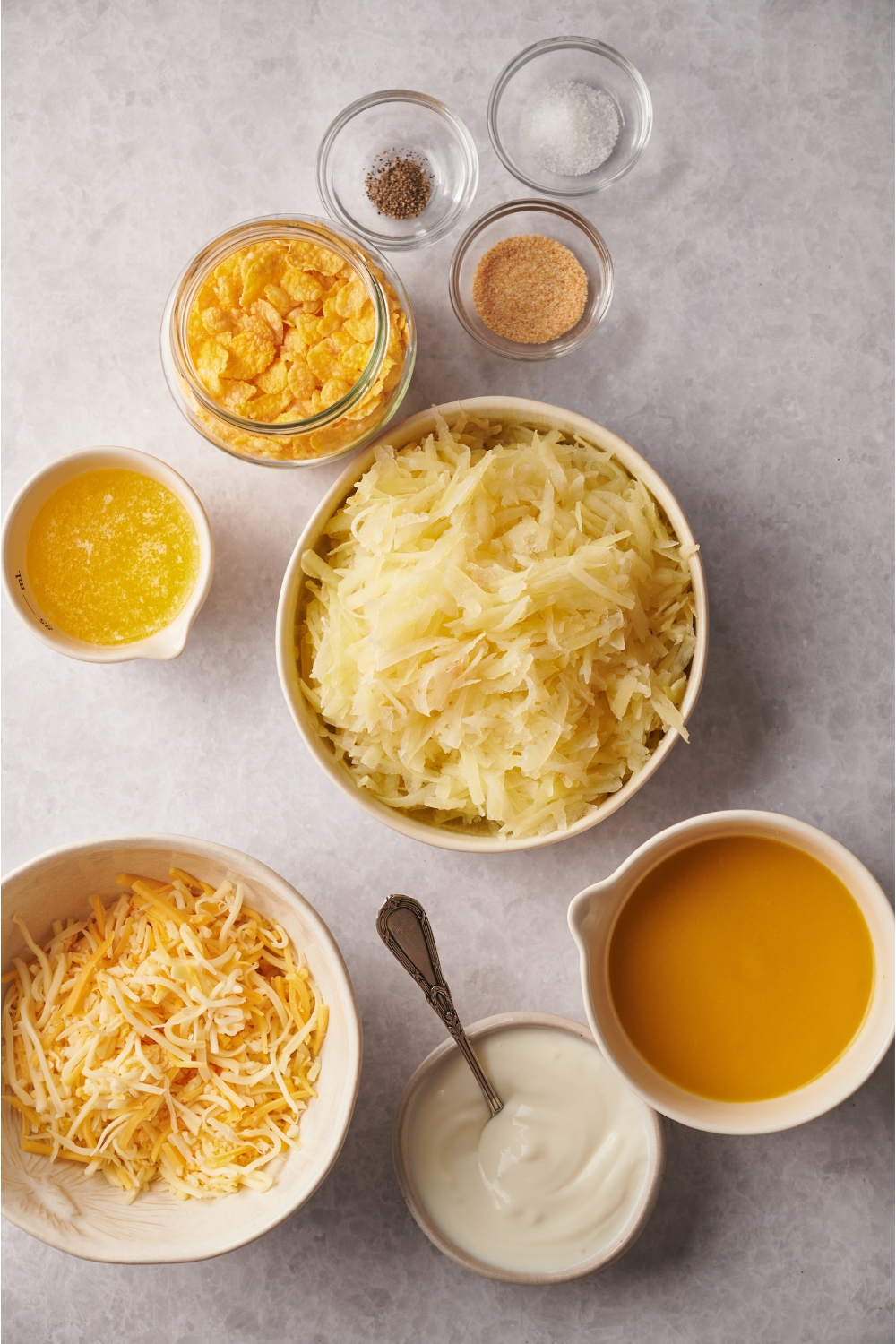 A countertop with hashbrowns, shredded cheese, broth, condensed soup, melted butter, corn flakes and seasonings in various bowls.