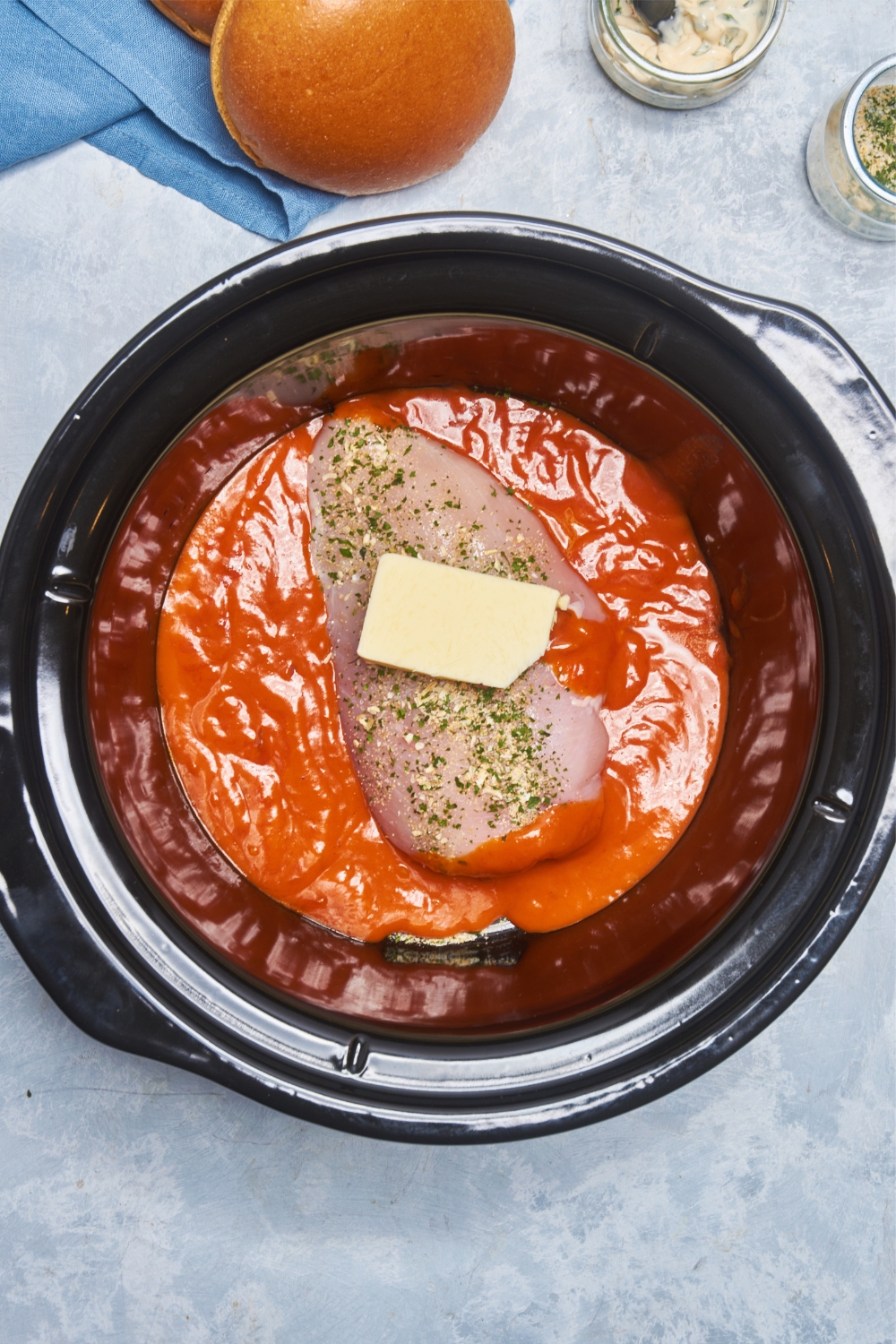 A chicken breast with ranch seasoning and a pat of butter in a crock pot filled with buffalo sauce.