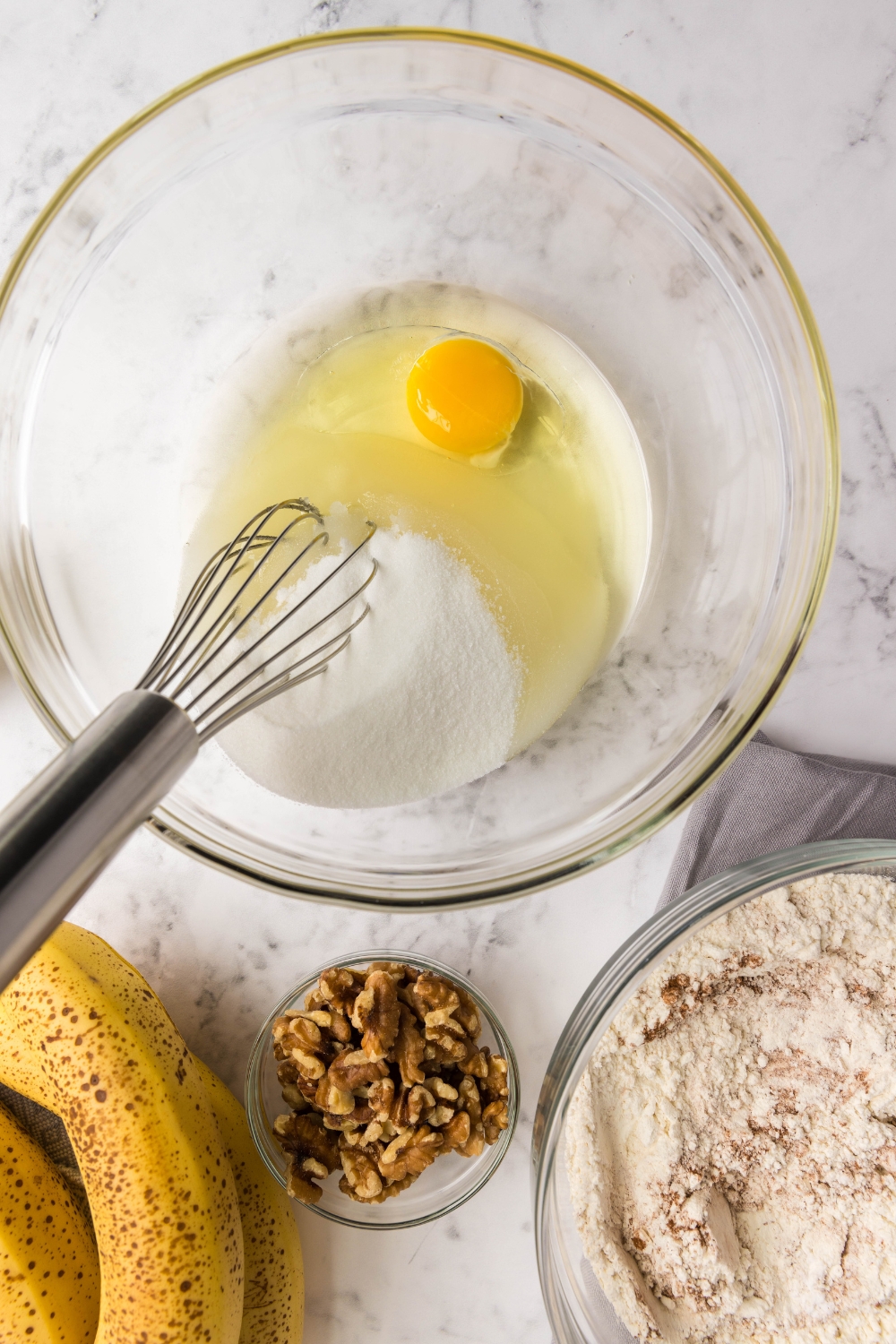 A mixing bowl with a cracked egg, oil. and sugar. A whisk is sitting in the bowl.