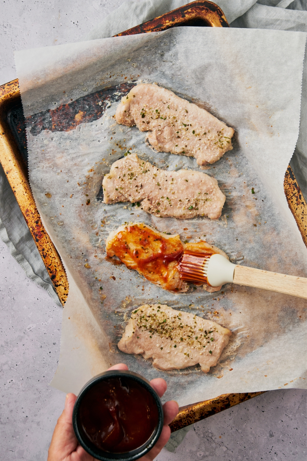 BBQ sauce being brushed onto seasoned pork chops on a parchment paper lined baking sheet using a pastry brush.