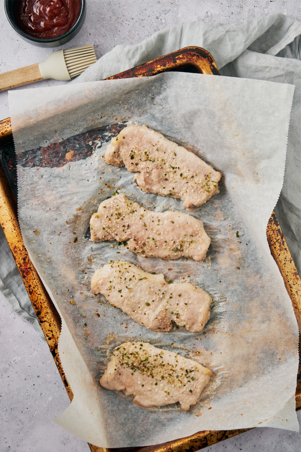 Four seasoned pork chops on a parchment paper lined baking sheet next to a bowl of sauce and a pastry brush.