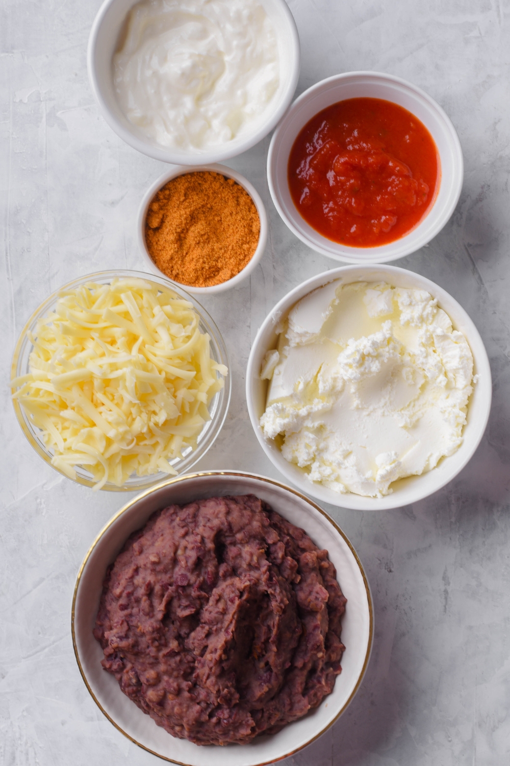 A countertop with cream cheese, sour cream, salsa, taco seasoning, and refried beans in separate bowls.