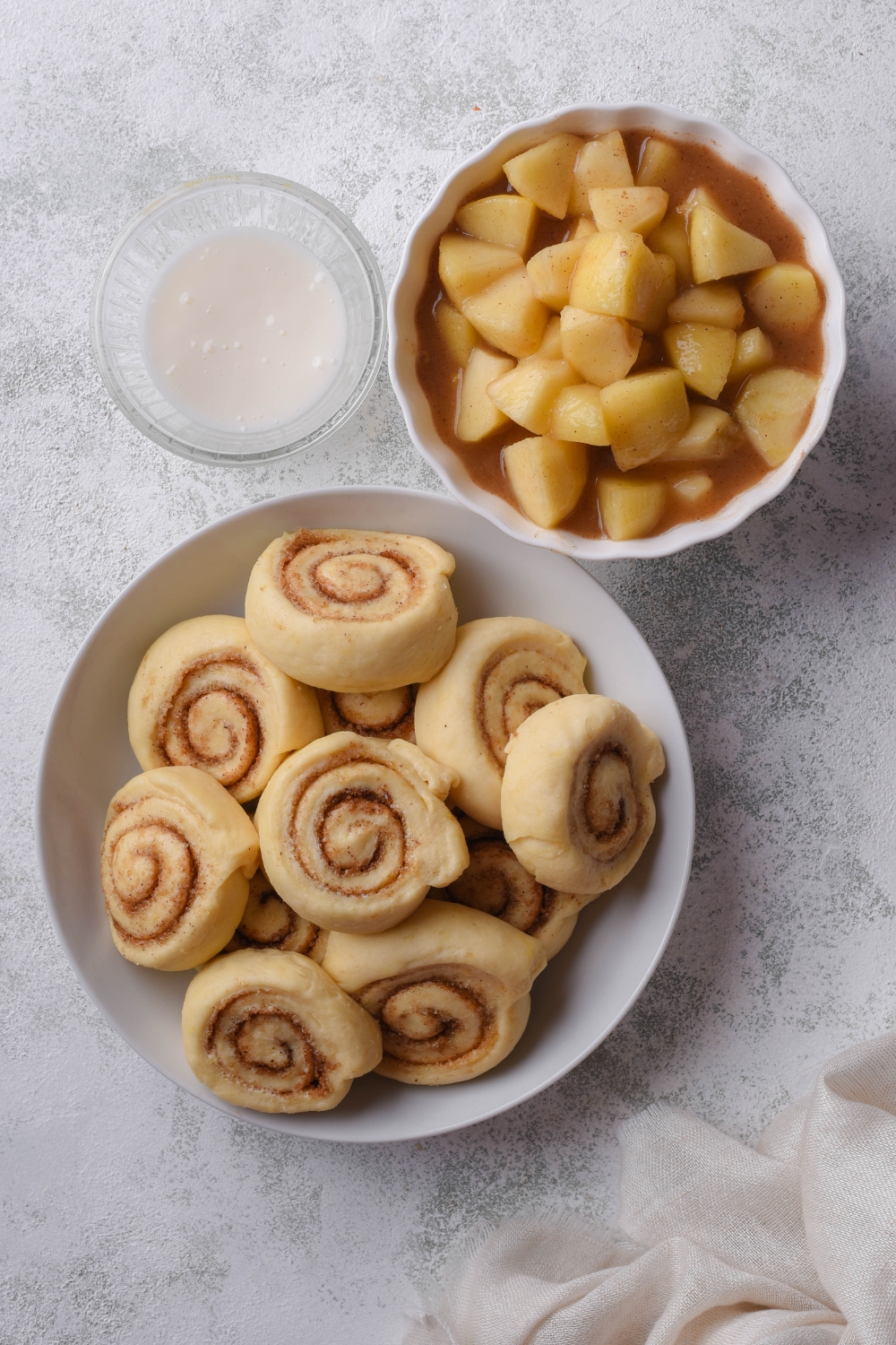 A countertop with various bowls with cinnamon roll dough, apple pie filling, and icing.