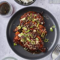 Top view of Korean pork chops garnished with chopped green onions and sesame seeds on a black plate.