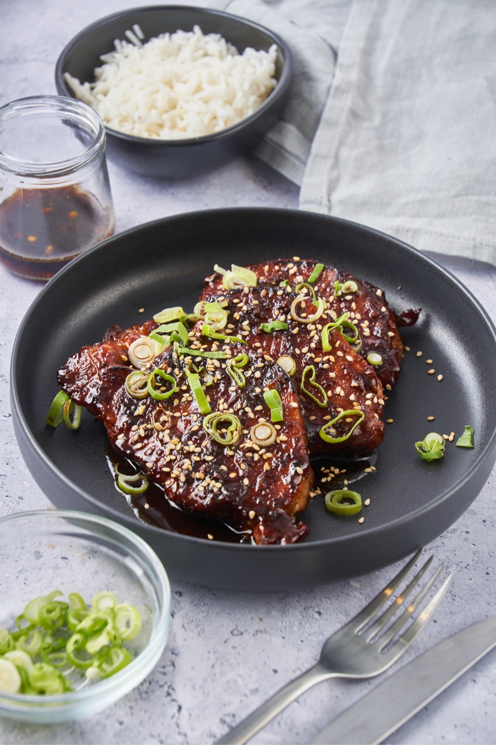 Korean pork chops garnished with chopped green onions and sesame seeds in a black plate served with a side of white rice.