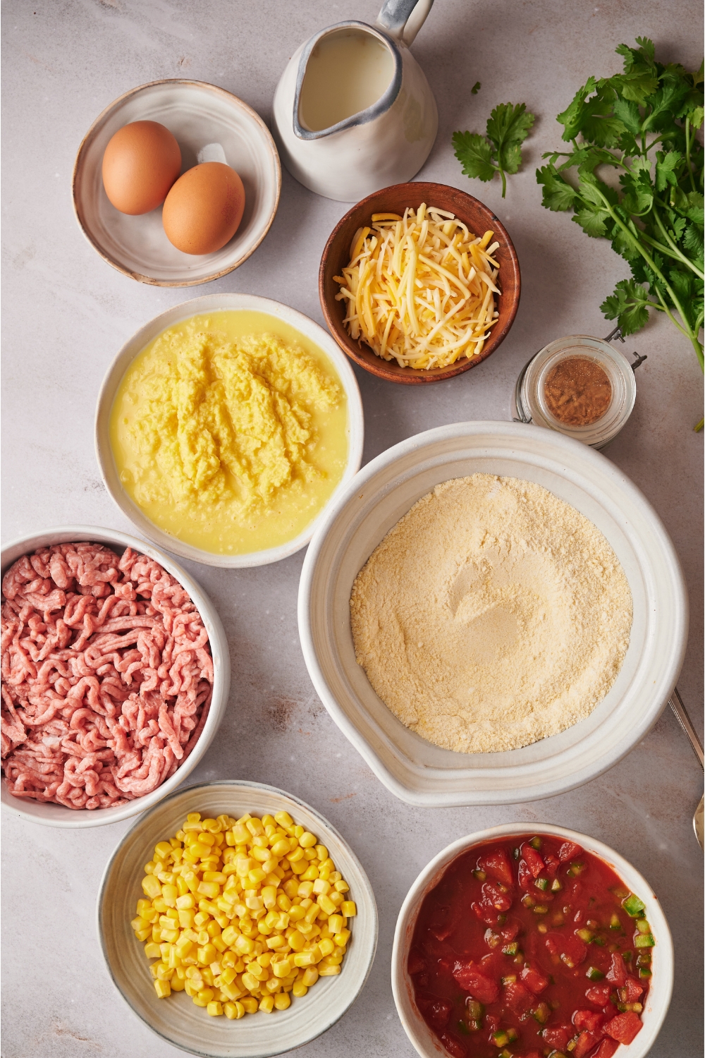 A bowl with two eggs, a bowl of shredded cheese, a bowl of jiffy cornbread mix, a bowl of ground beef, a bowl of corn, a bowl of creamed corn, and a bowl of crushed tomatoes all on a grey counter.