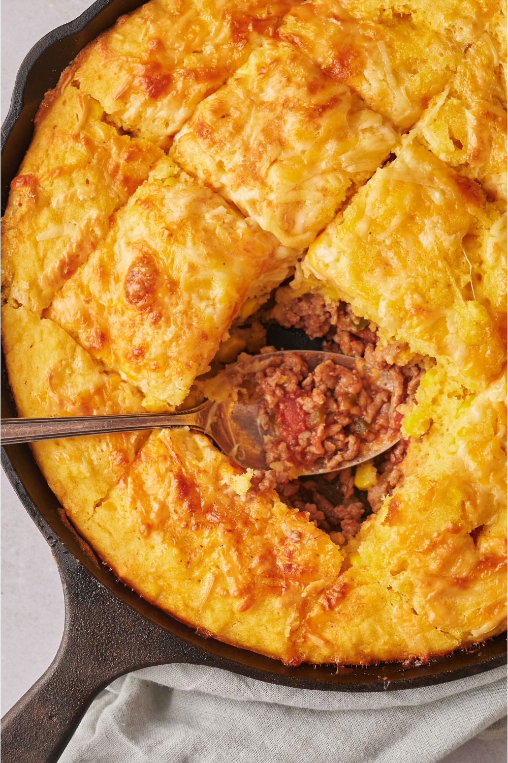Part of a cast iron skillet with mexican cornbread casserole in it. One slice is missing and there is a spoon in it with ground beef.