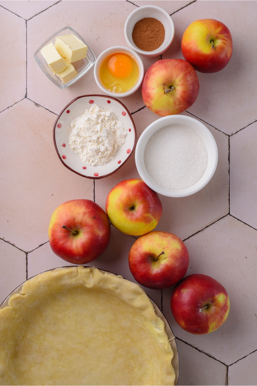A countertop with apples, flour, sugar, egg, cinnamon, butter, and pie crust in various bowls and containers.