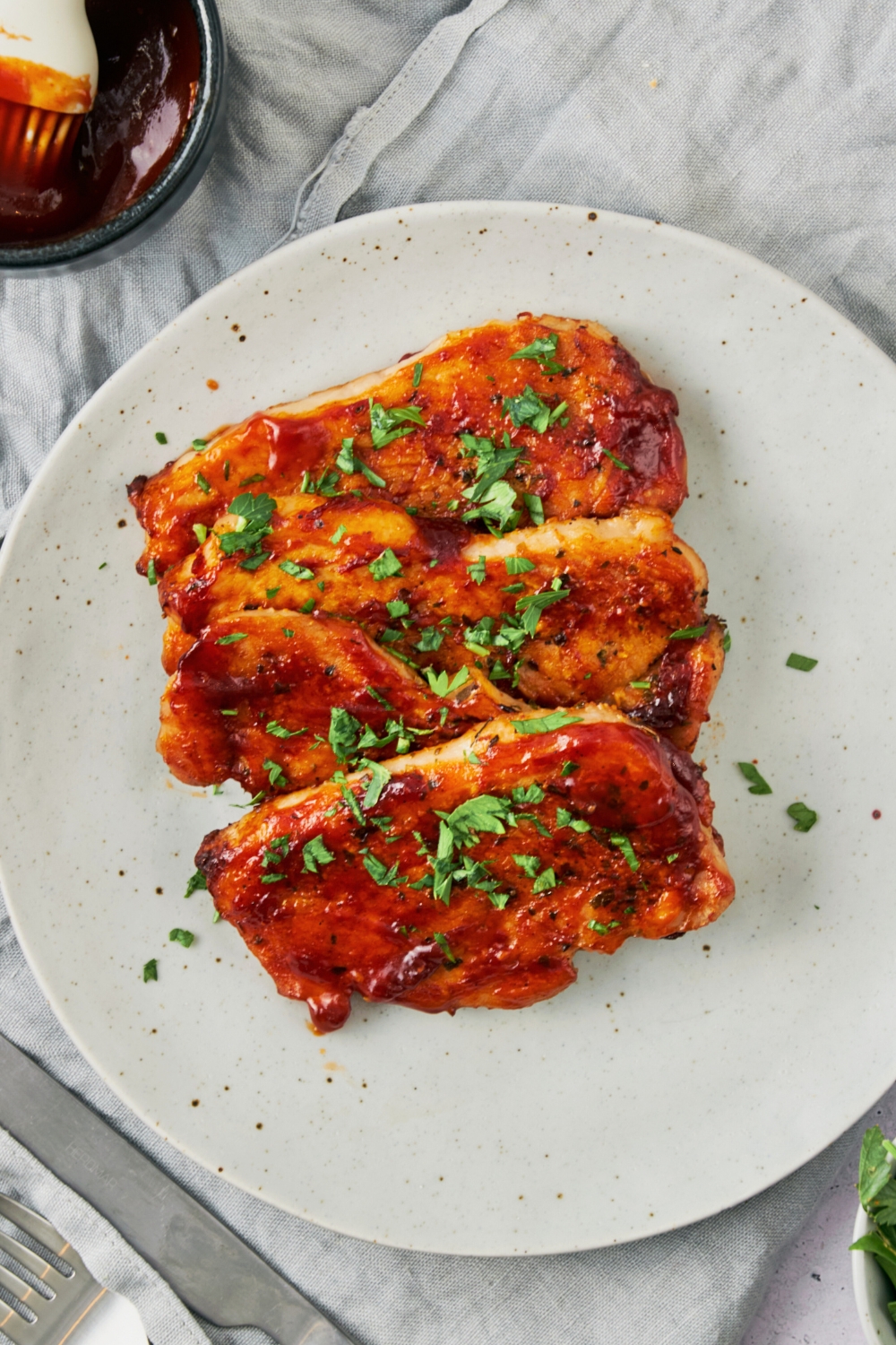 Three bbq pork chops overlapping one another on a white plate on a grey tablecloth.