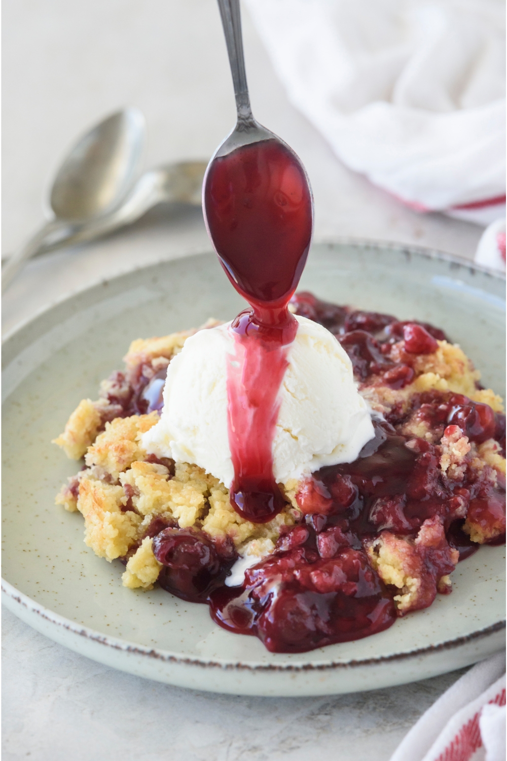 A plate with cherry dump cake topped with a scoop of vanilla ice cream. A spoon id drizzling some cherry juice on the ice cream.