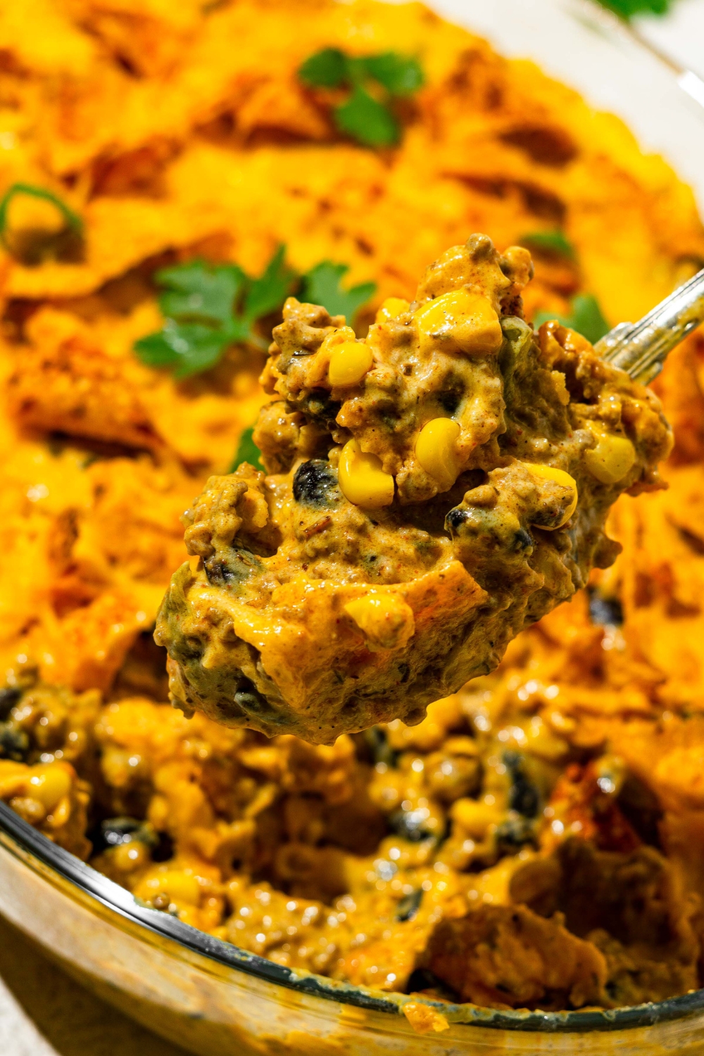 A spoon with dorito taco casserole on it being held above the casserole in a baking dish.