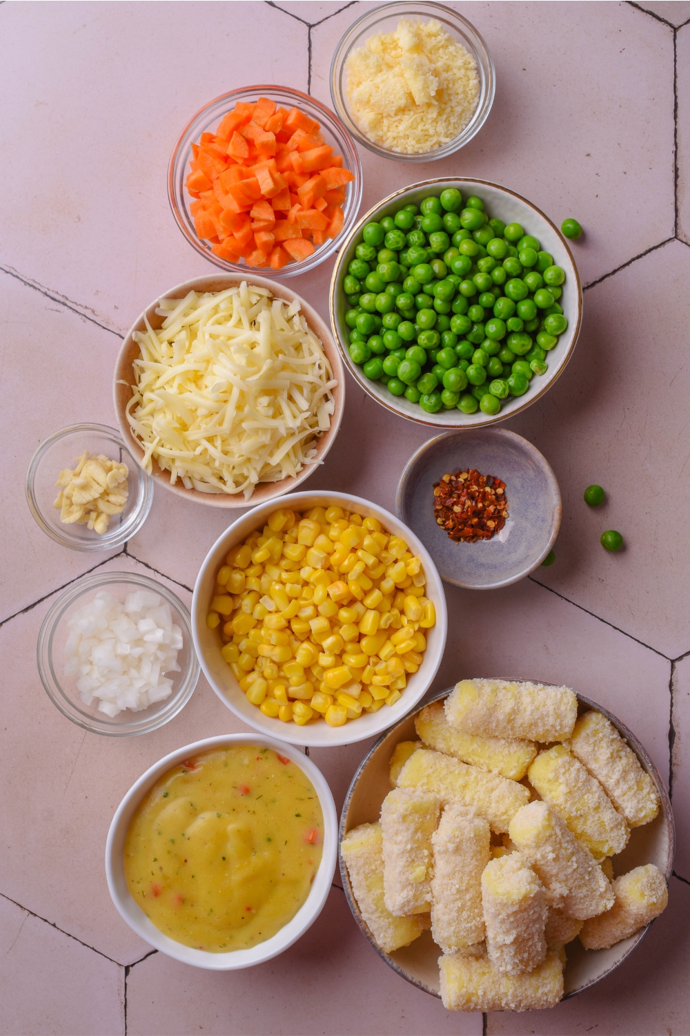 A countertop with multiple bowls containing peas, carrots, shredded cheese, red pepper flakes, corn, garlic, onion, condensed soup, and tater tots.