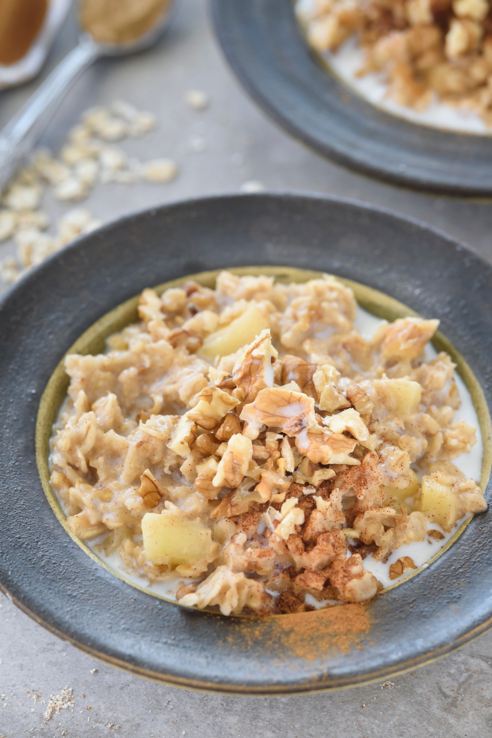 A bowl if oatmeal with milk covered in diced apples, chopped nuts, and ground cinnamon. In the background is a second bowl of oatmeal.