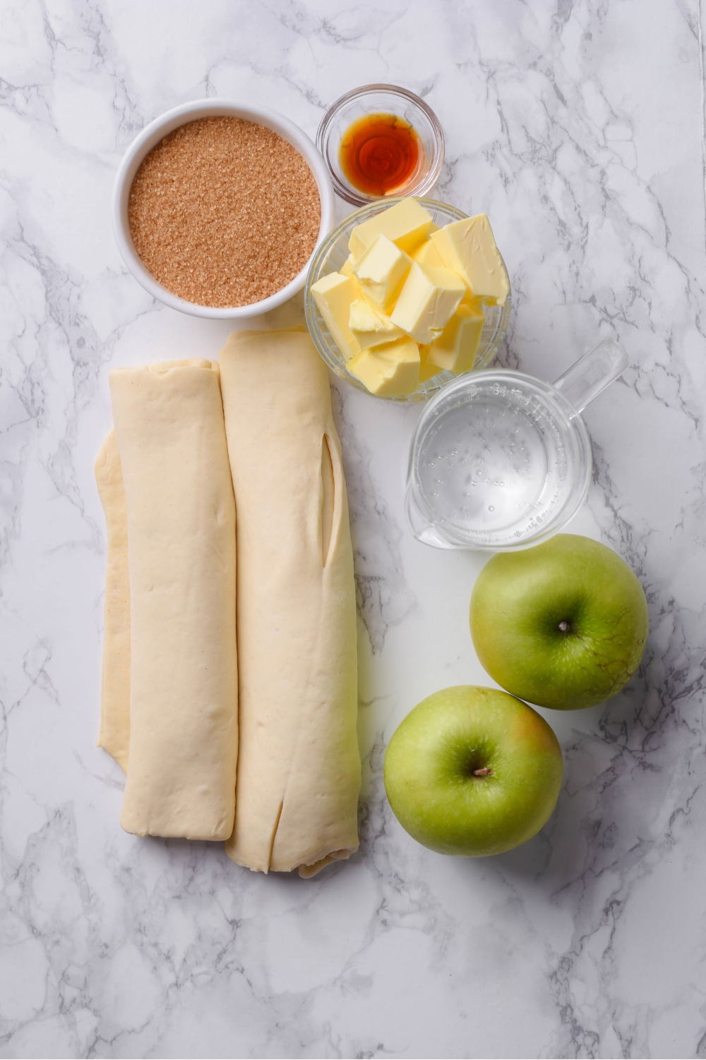 A countertop with multiple bowls with butter, lemon lime soda, brown sugar, vanilla extract, cinnamon, crescent roll dough, and two apples.