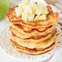 A tall stack of golden brown pancakes topped with diced apples.