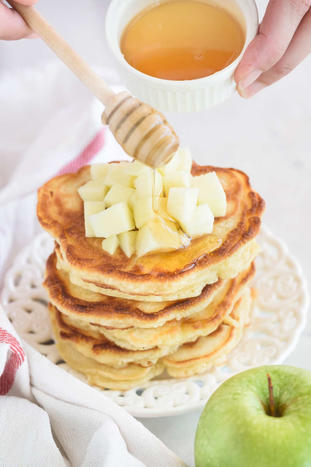 A tall stack of pancakes topped with diced apples and honey being drizzled on top of the pancakes.