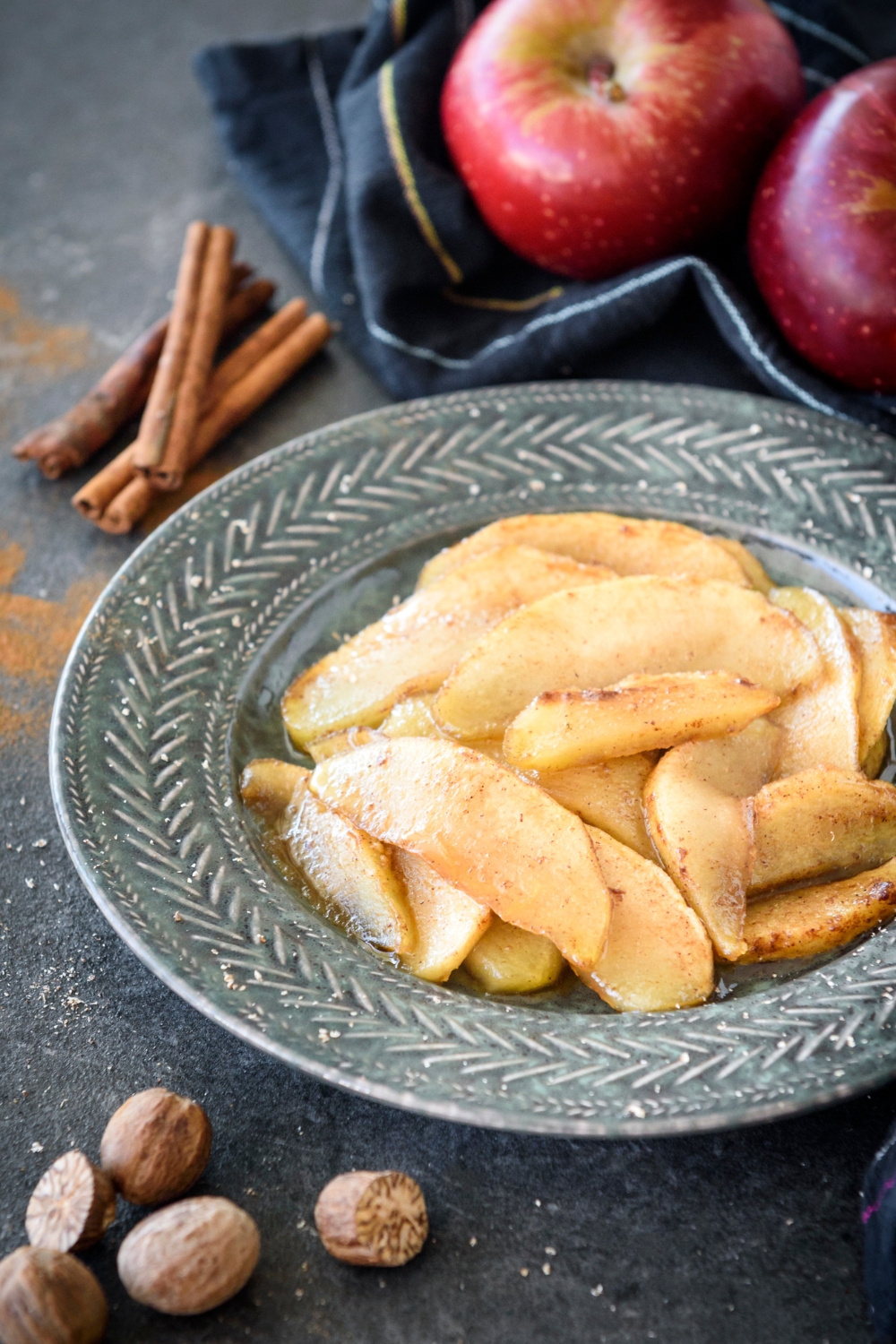 A bowl of baked apple slices coated in spices surrounded by cinnamon, nutmeg, and apples.