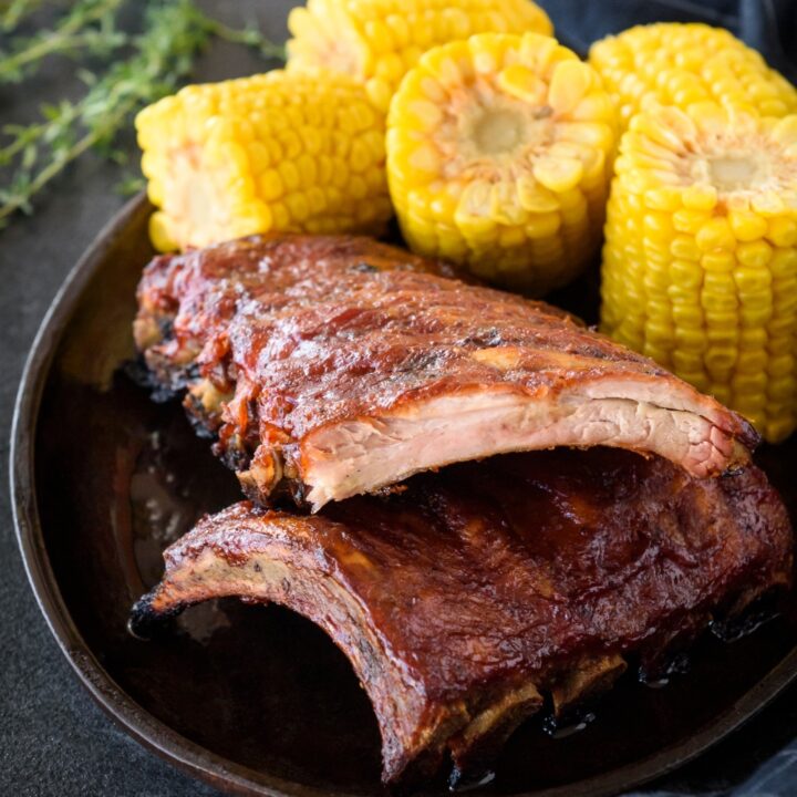 Two racks of ribs stacked on top of each other, each covered with barbecue sauce and a side of sliced corn cobs.